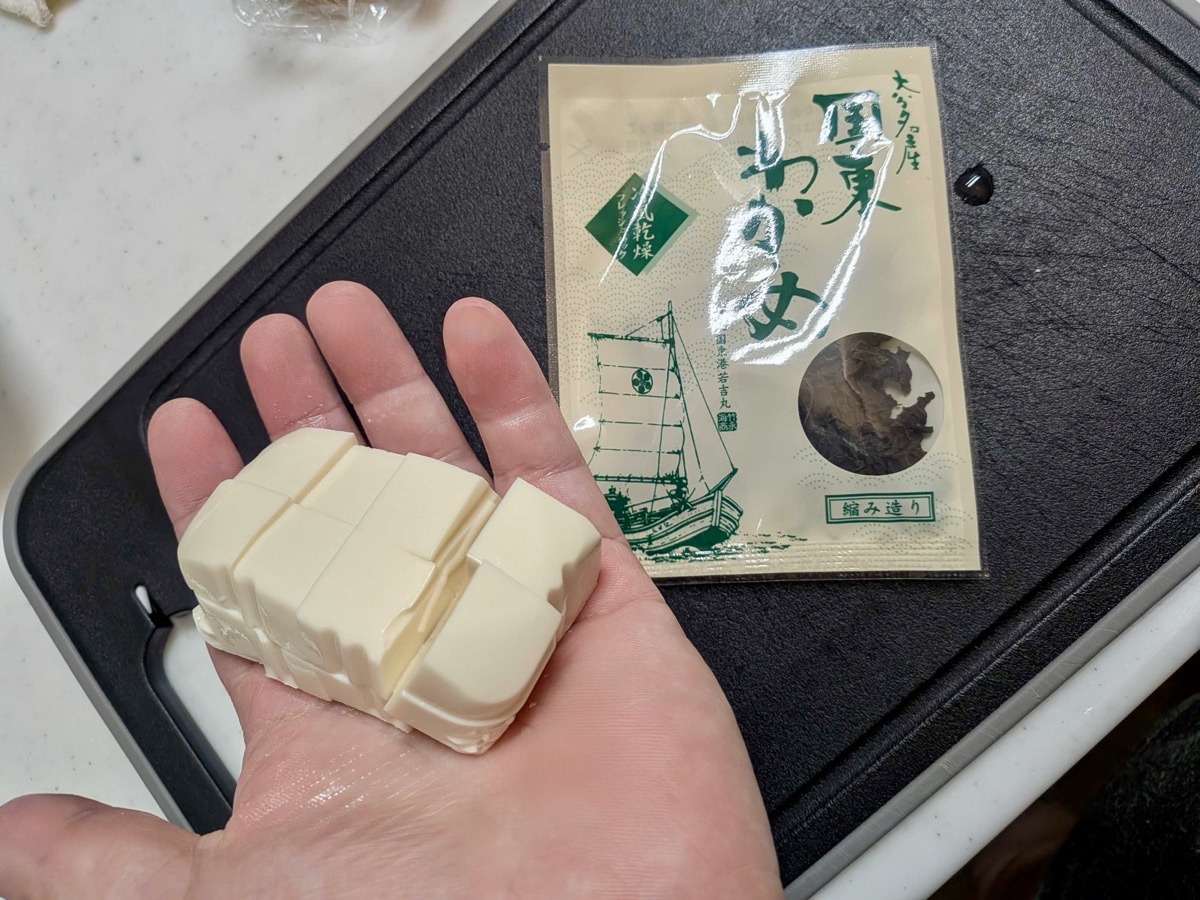 Cut tofu held in hand next to a dried wakame packet on a cutting board