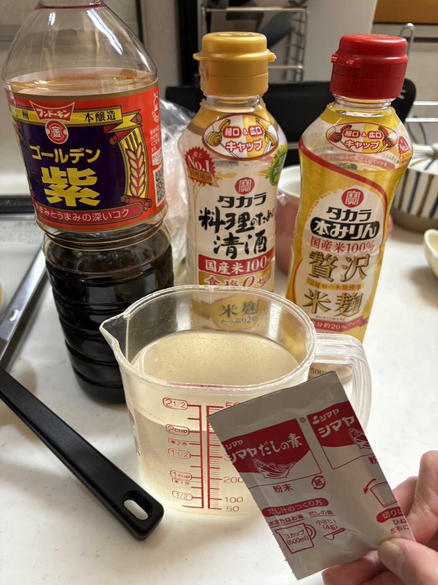 Japanese cooking seasonings including soy sauce, cooking sake, mirin, and dashi powder packet arranged on a kitchen counter with a measuring cup