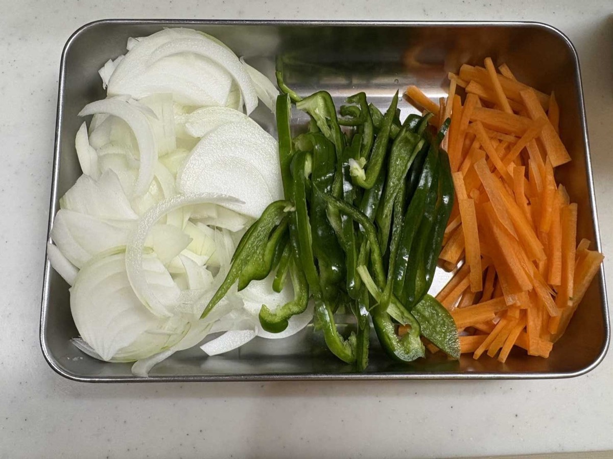 Thinly sliced onion, julienned carrot matchsticks, and green pepper strips arranged separately in a stainless steel tray, ready for the stir-fry