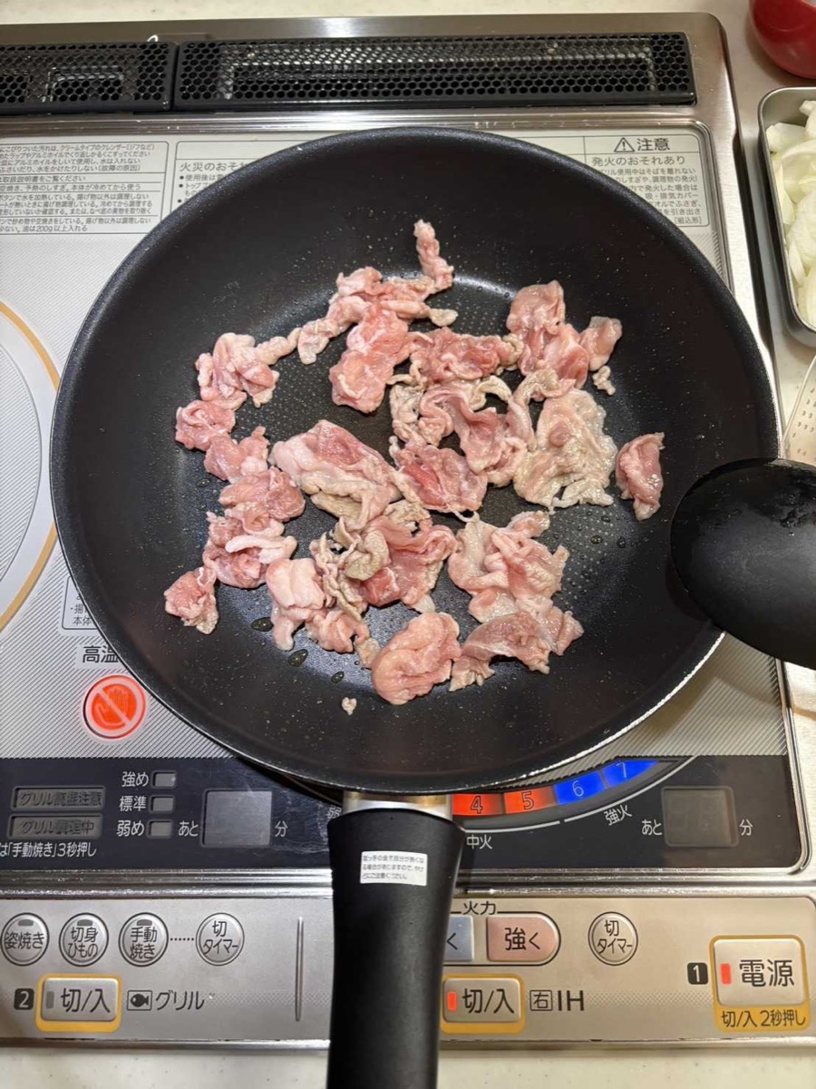 Thinly sliced pork dusted with potato starch stir-frying in sesame oil in a black frying pan on an IH cooktop, starting to turn from pink to pale brown