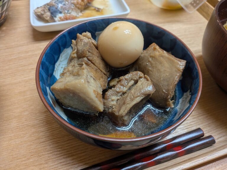 A finished bowl of buta no kakuni — Japanese braised pork belly — served in a blue and white Japanese ceramic bowl with a glossy soy-coloured broth, a soft-boiled egg, and chopsticks on a wooden tray