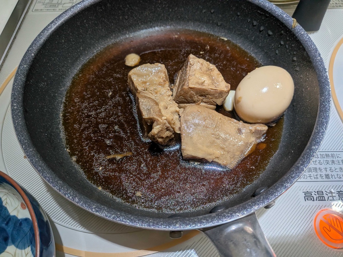 Three cubes of finished braised pork belly kakuni glossy with sauce, plated alongside a deeply tea-coloured soft-boiled egg in a frying pan