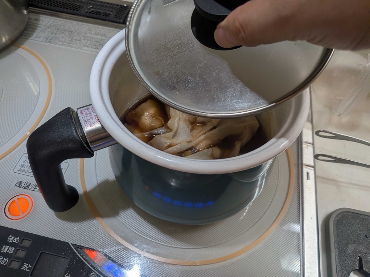 A sheet of parchment paper being placed directly on the surface of the simmering kakuni braising liquid — a parchment otoshibuta for even cooking