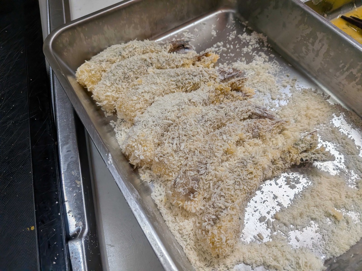 Shrimp fully coated in panko breadcrumbs, resting in a metal tray ready to deep fry — alongside a few other panko-breaded items being fried that night