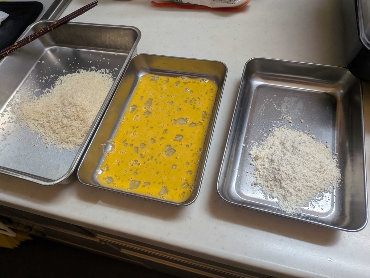 Three metal trays set up as a breading station — panko on the left, beaten egg in the middle, all-purpose flour on the right