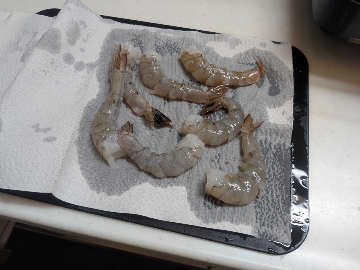 Cleaned and rinsed shrimp laid out on paper towels on a black tray, being patted dry before breading