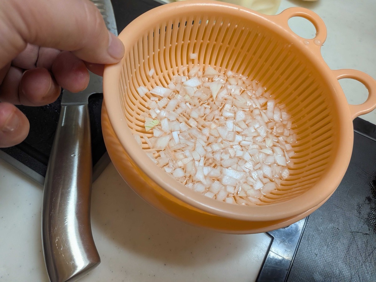 Finely minced onion in a peach-coloured colander being soaked in cold water to remove the sharp pungency