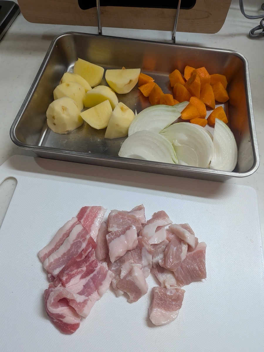 Prepared curry ingredients: pork belly pieces on a cutting board with potato, carrot, and onion cut into chunks in a stainless steel tray