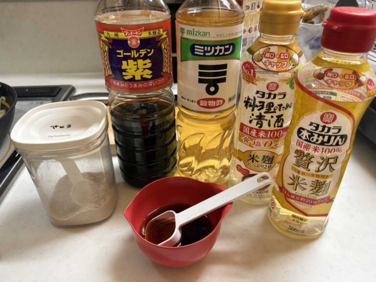 A row of essential Japanese pantry seasonings on a kitchen counter — soy sauce, rice vinegar, cooking sake, mirin, and a container of sugar — the foundation of Japanese home cooking