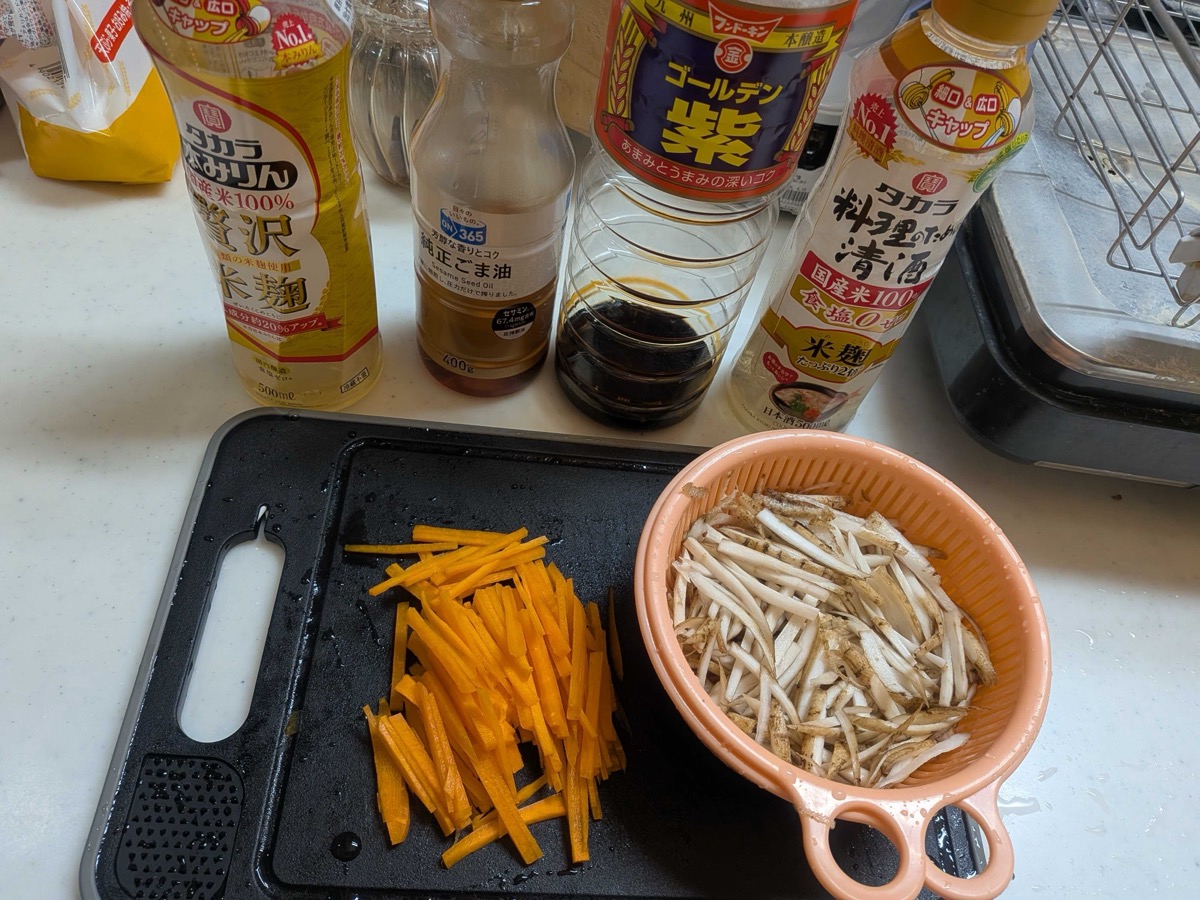 Julienned burdock root and carrot on a cutting board alongside Japanese seasonings including mirin, sesame oil, soy sauce, and cooking sake