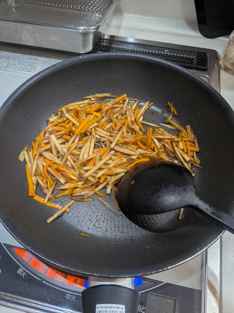 Kinpira gobo nearly finished cooking in a pan, with the sauce almost fully reduced and the vegetables deeply glazed