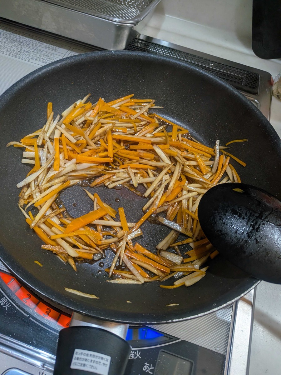 Burdock root and carrot simmering with soy sauce and mirin glaze in a frying pan, with the sauce beginning to reduce