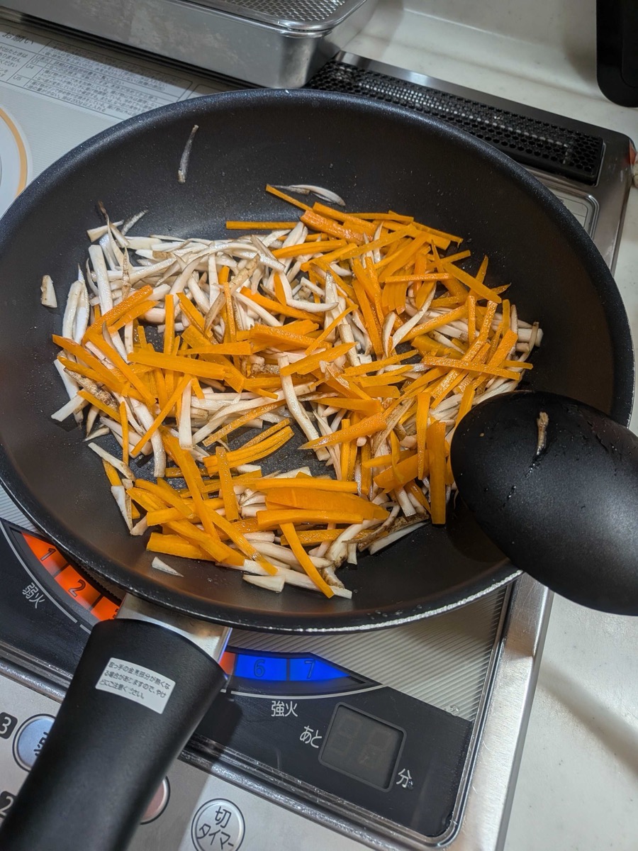 Julienned burdock root and carrot stir-frying in sesame oil in a black frying pan on an IH cooktop