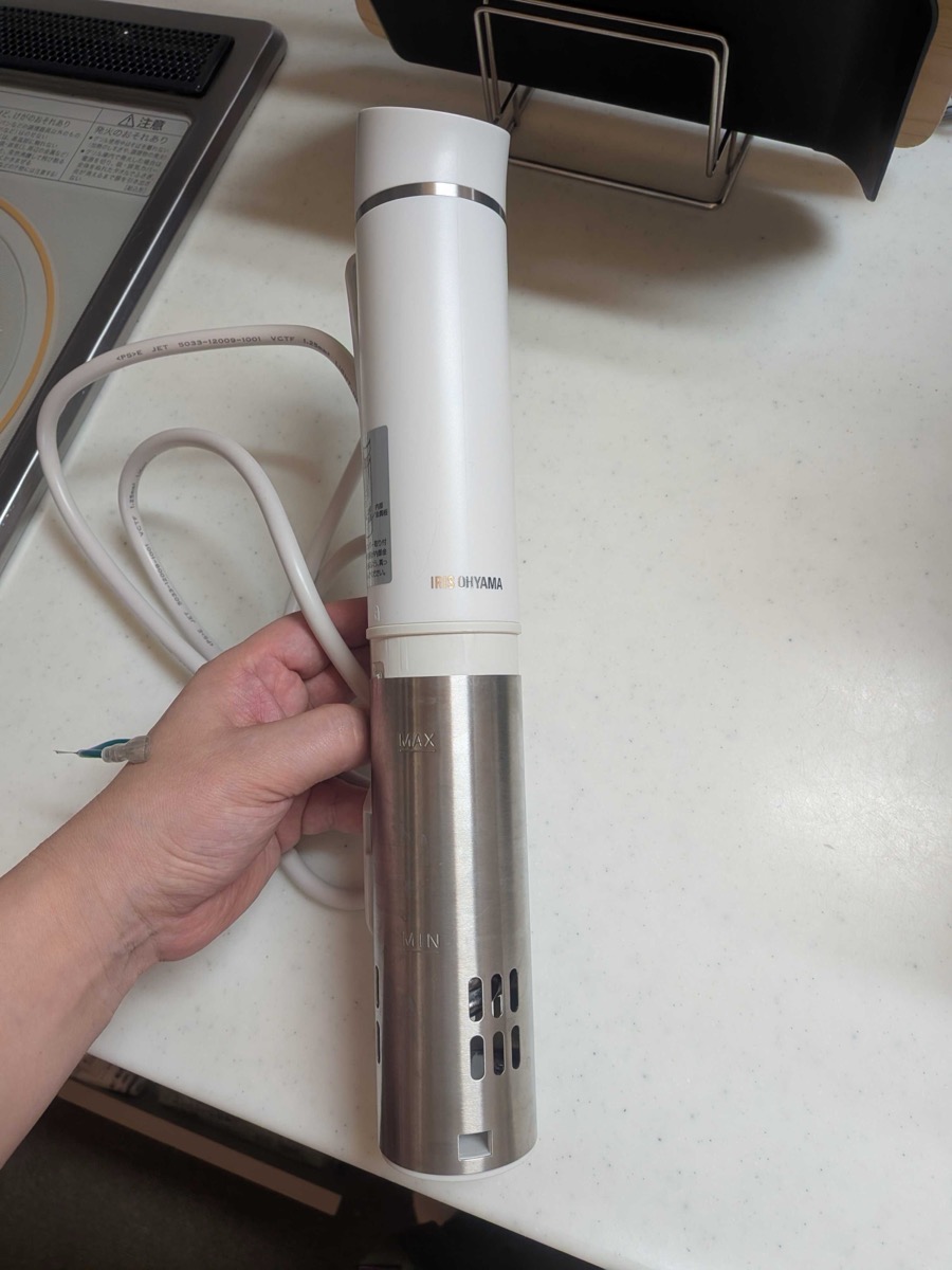 An Iris Ohyama sous vide immersion circulator held in hand on a kitchen counter, used for cooking tonkatsu pork at 63°C for 90 minutes