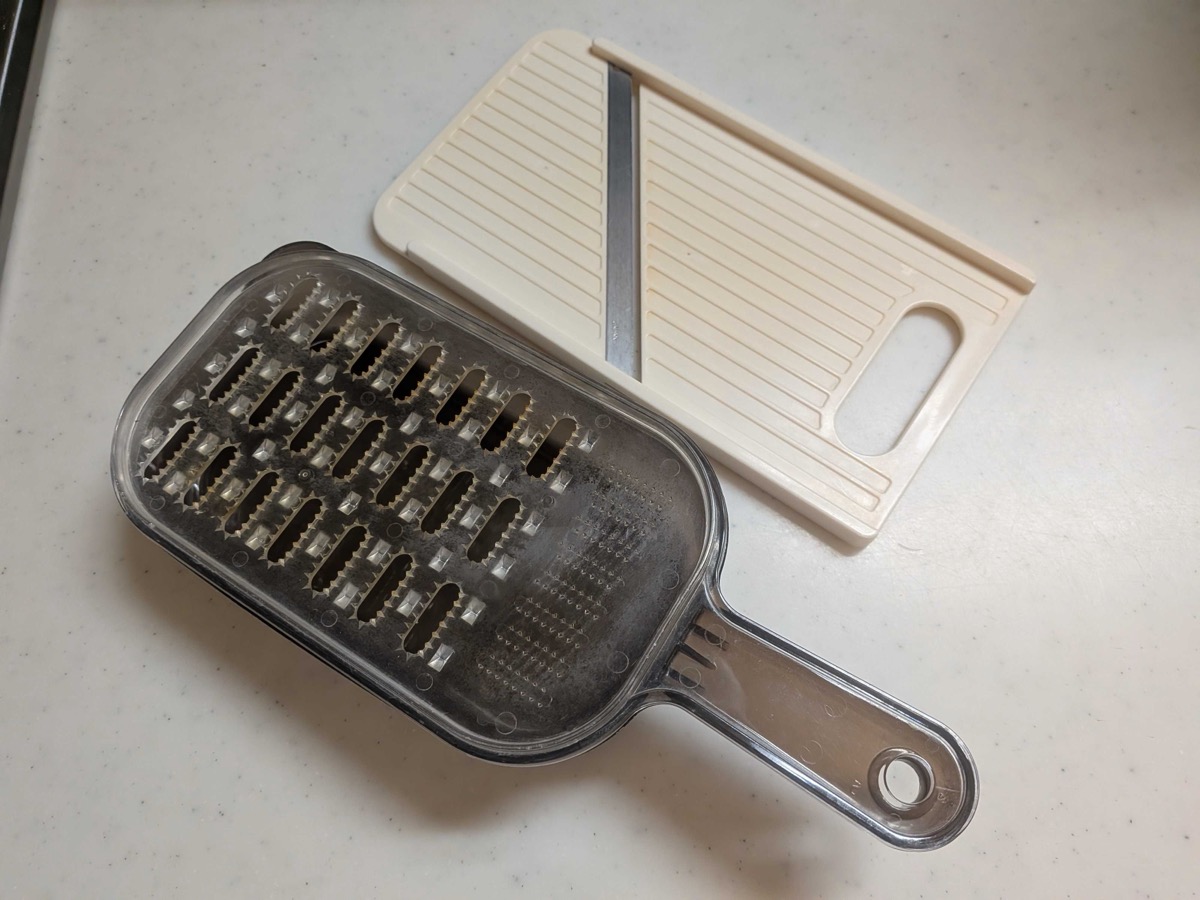 A Japanese mandoline slicer (white plastic) next to a coarse oni-oroshi style grater (clear plastic with metal teeth) on a kitchen counter — two essential Japanese vegetable prep tools