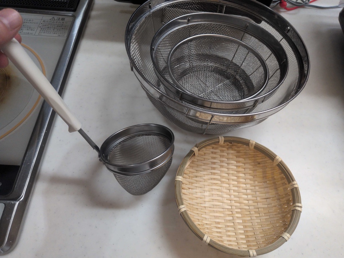 A collection of Japanese kitchen strainers — three nested stainless steel mesh zaru, a small mesh skimmer, and a traditional woven bamboo zaru — all essential for Japanese cooking prep and serving