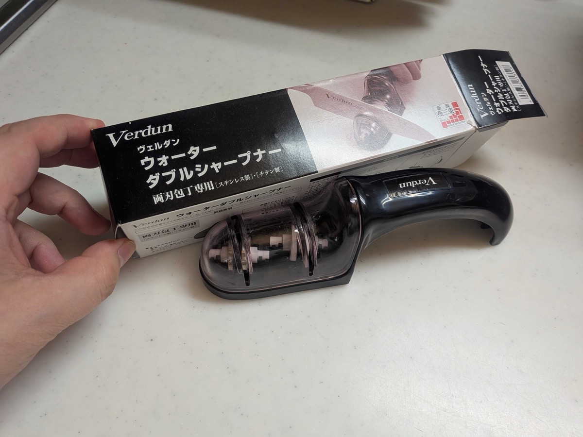 A Verdun water double sharpener held in hand next to its packaging — a Japanese pull-through knife sharpener for double-bevel stainless and titanium blades, made in Niigata Prefecture