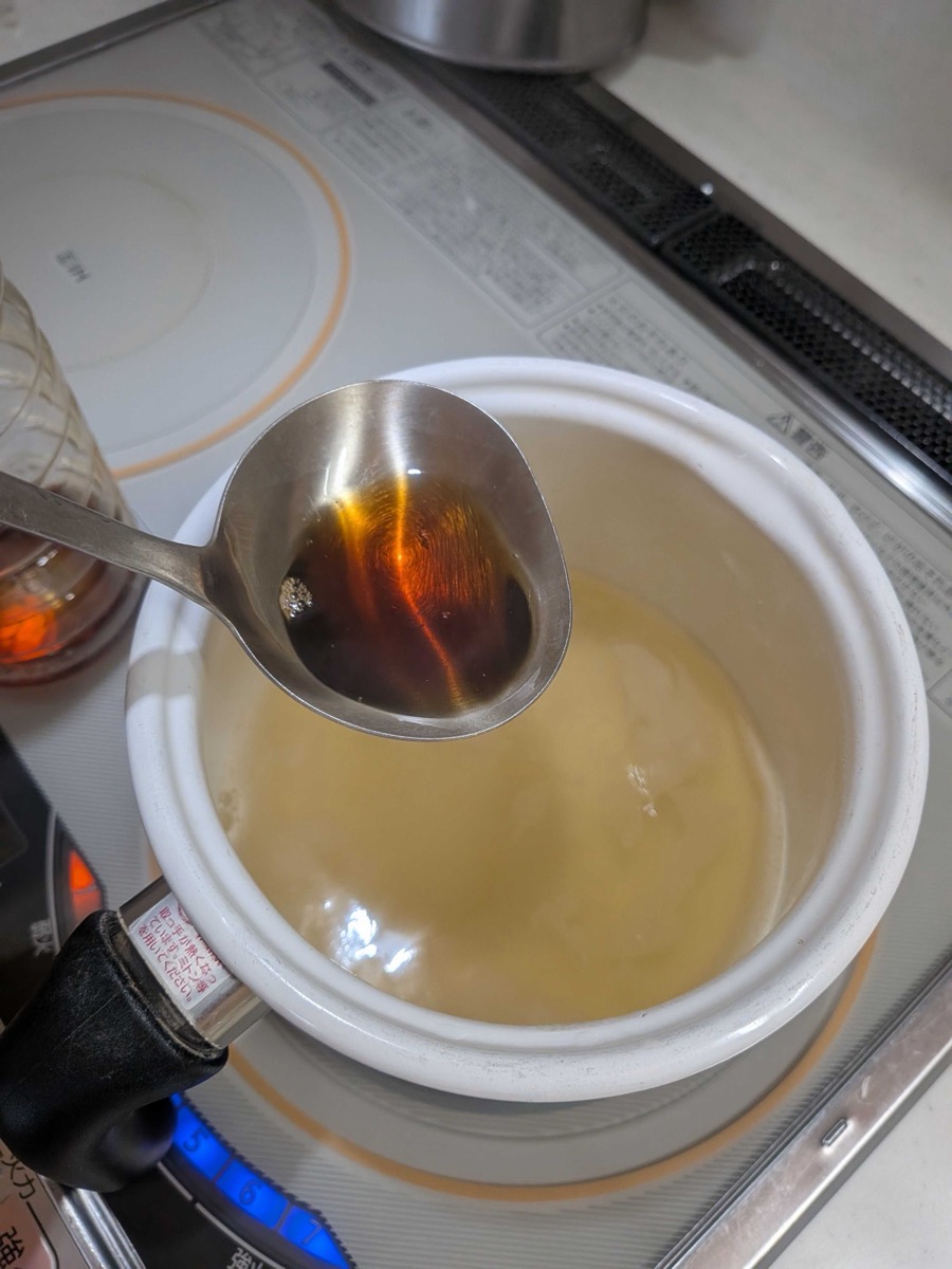 A spoonful of dark usukuchi soy sauce being added to a pot of clear golden dashi broth on an IH cooktop