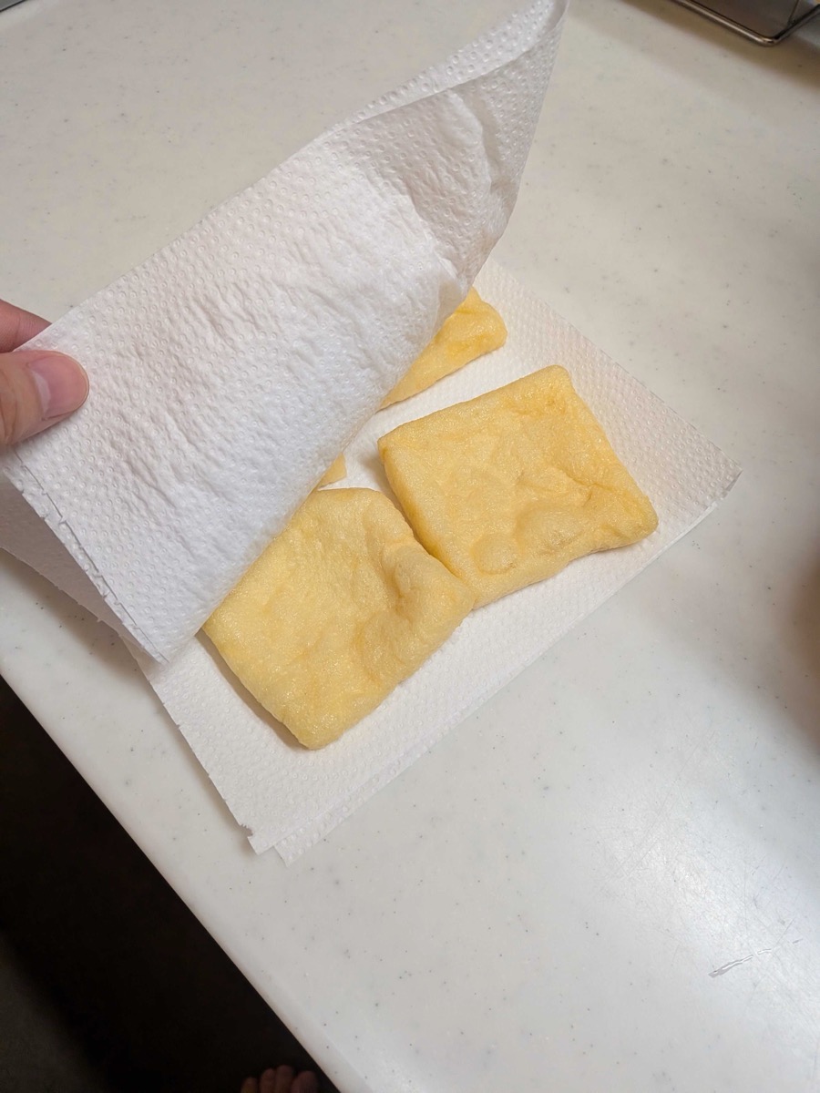 Aburaage tofu pouches being pressed between paper towels to remove excess water after the abura-nuki rinse