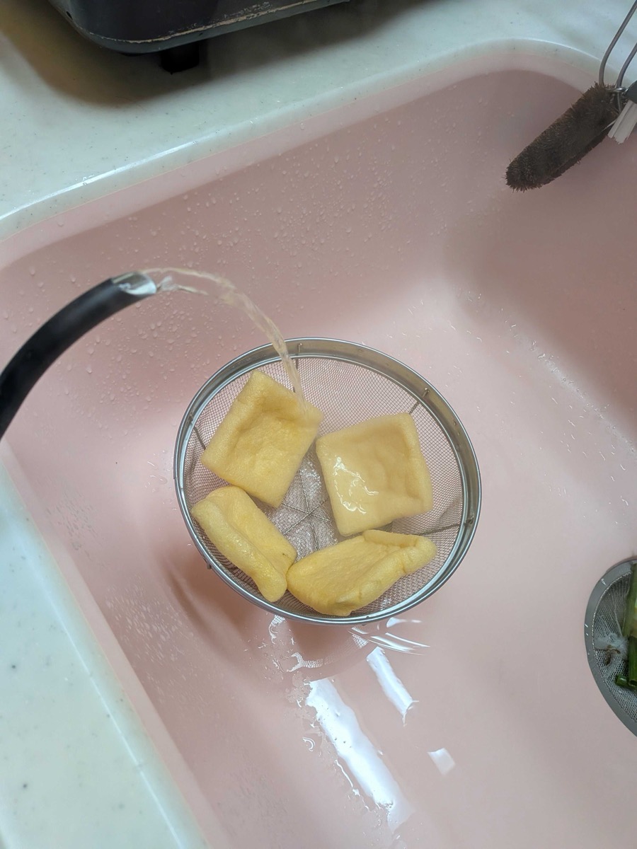 Four square aburaage tofu pouches in a mesh strainer being rinsed with hot water from the tap to remove surface oil — abura-nuki step