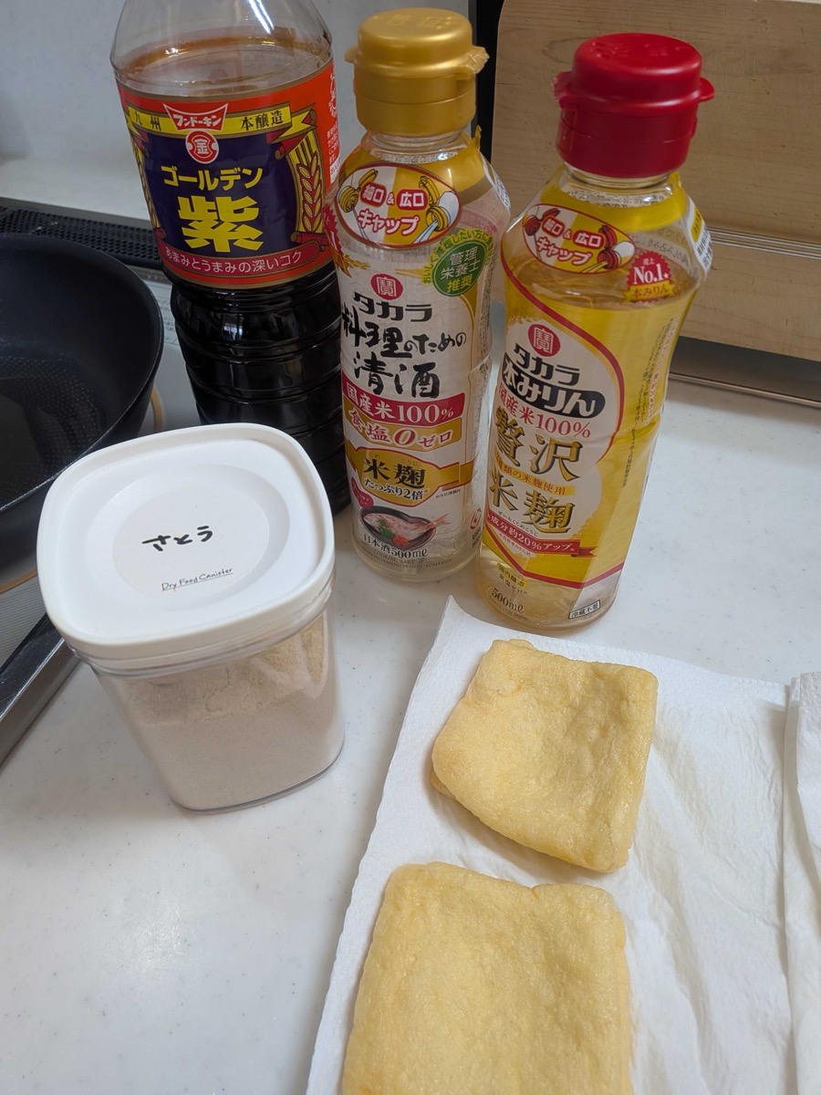 Japanese seasonings for kitsune udon age — soy sauce, sake, mirin, and a sugar container — set out on a counter alongside the prepared aburaage on paper towels
