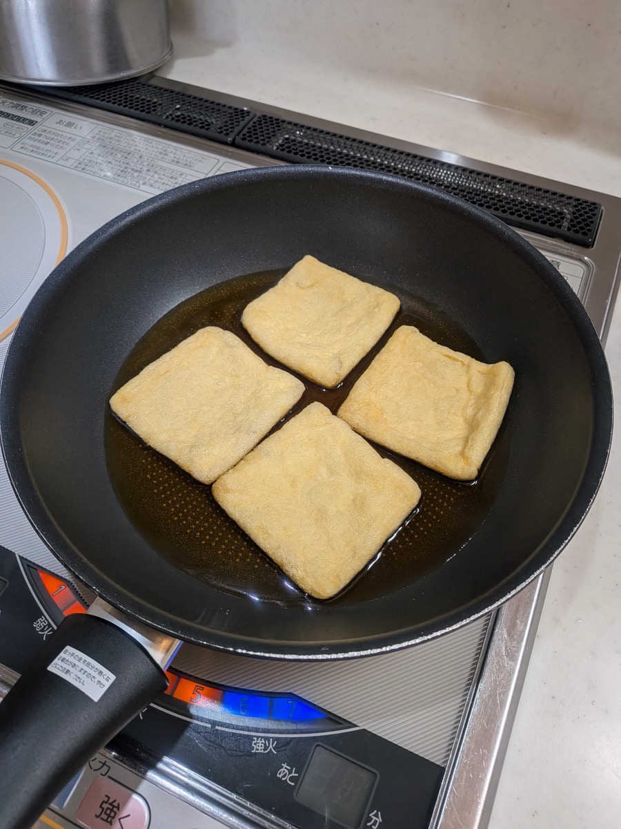 Four square aburaage tofu pouches arranged in a single layer in a black frying pan with the seasoning broth, just starting to simmer on an IH cooktop