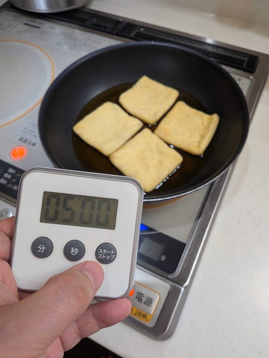 A kitchen timer set to 5 minutes held in front of a pan of simmering aburaage tofu pouches on an IH cooktop