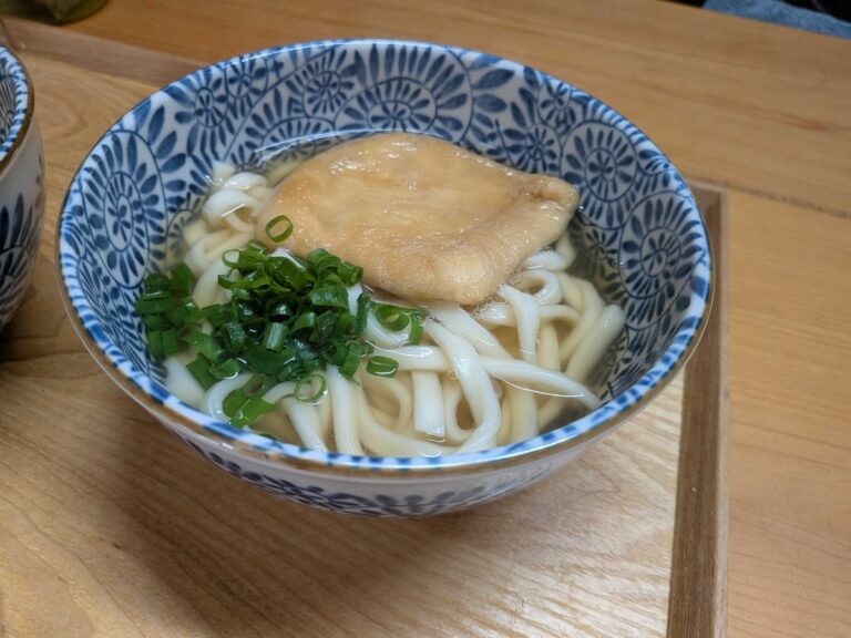A finished bowl of kitsune udon in a blue and white Japanese ceramic bowl, with golden broth, thick udon noodles, sweet simmered tofu pouch on top, and chopped green onion
