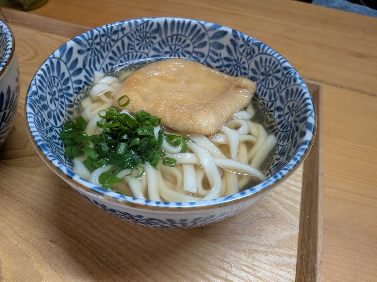A finished bowl of kitsune udon in a blue and white Japanese ceramic bowl, with golden broth, thick udon noodles, sweet simmered tofu pouch on top, and chopped green onion