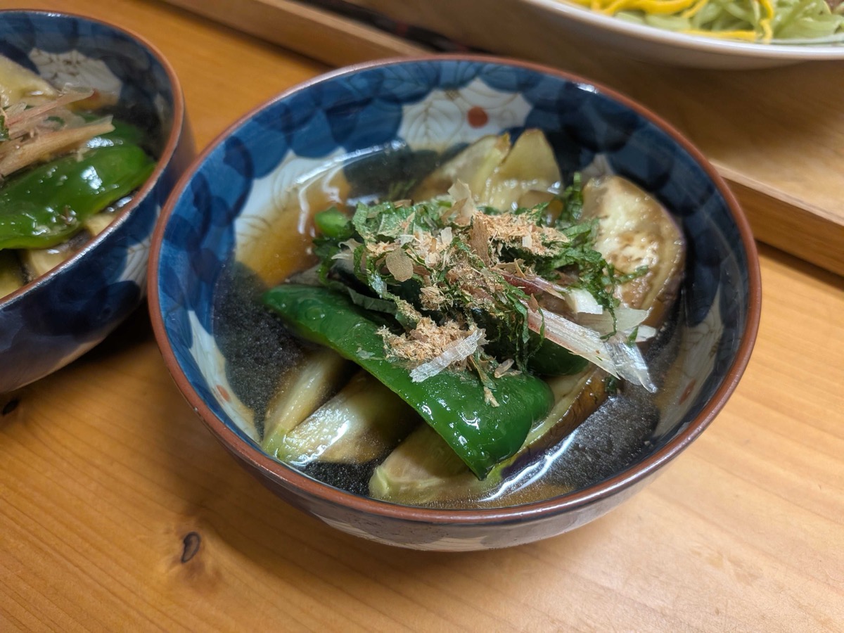 A finished bowl of nasu no yakibitashi — pan-fried Japanese eggplant and green pepper soaking in a dark dashi-soy tsuyu, topped with bonito flakes, shredded shiso, and myoga in a blue Japanese ceramic bowl