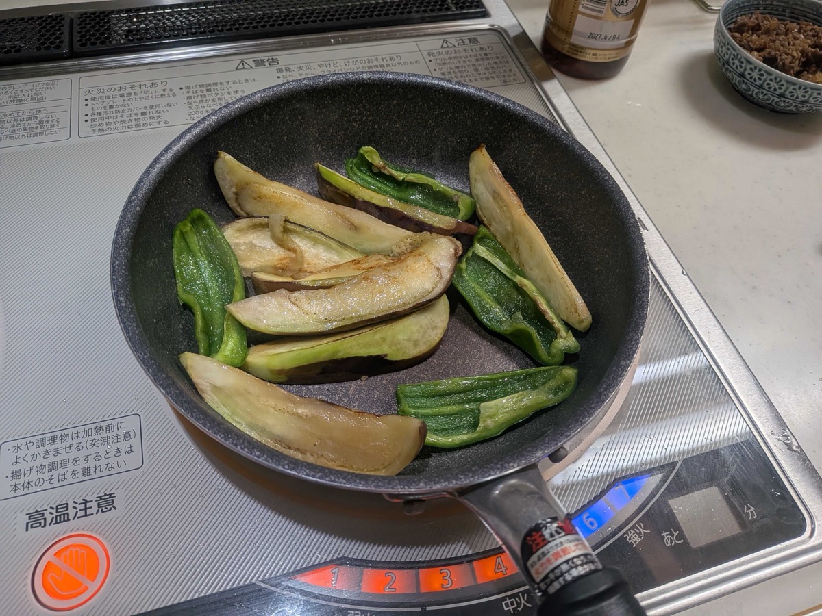 A close side view of the eggplant and green pepper finishing browning together in the frying pan, golden and glossy with sesame oil