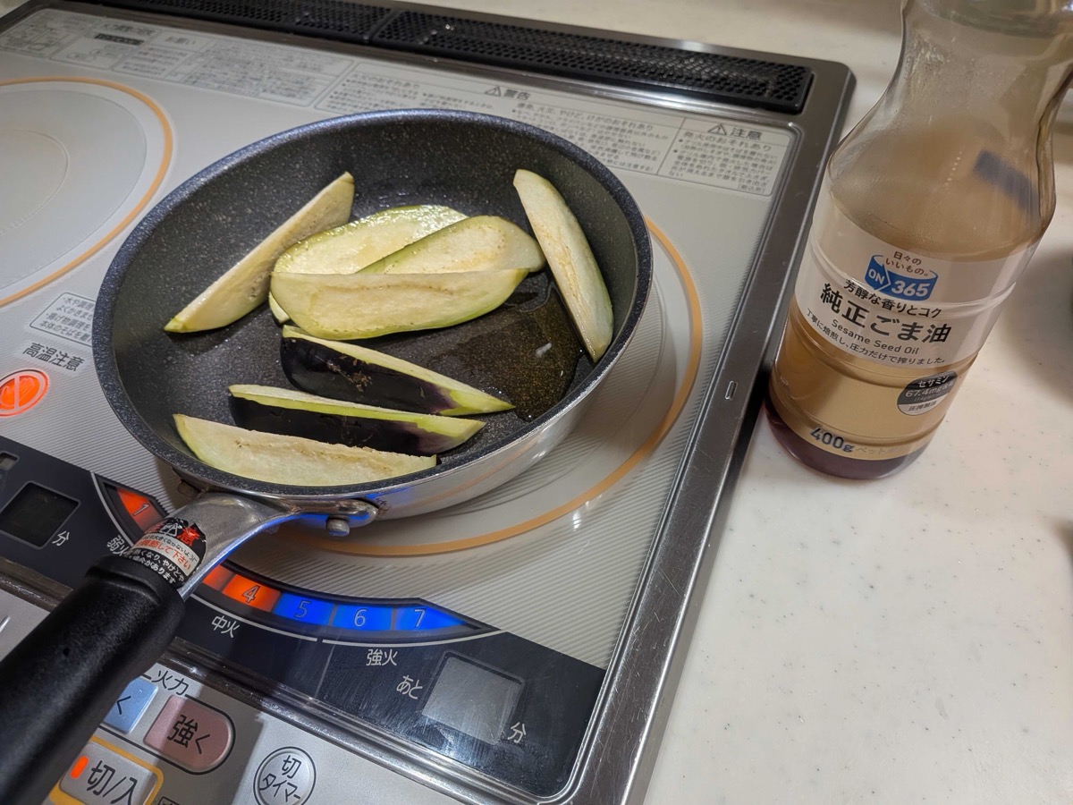 Eggplant wedges arranged cut-side up in a frying pan with sesame oil, with a bottle of pure sesame oil beside the cooktop