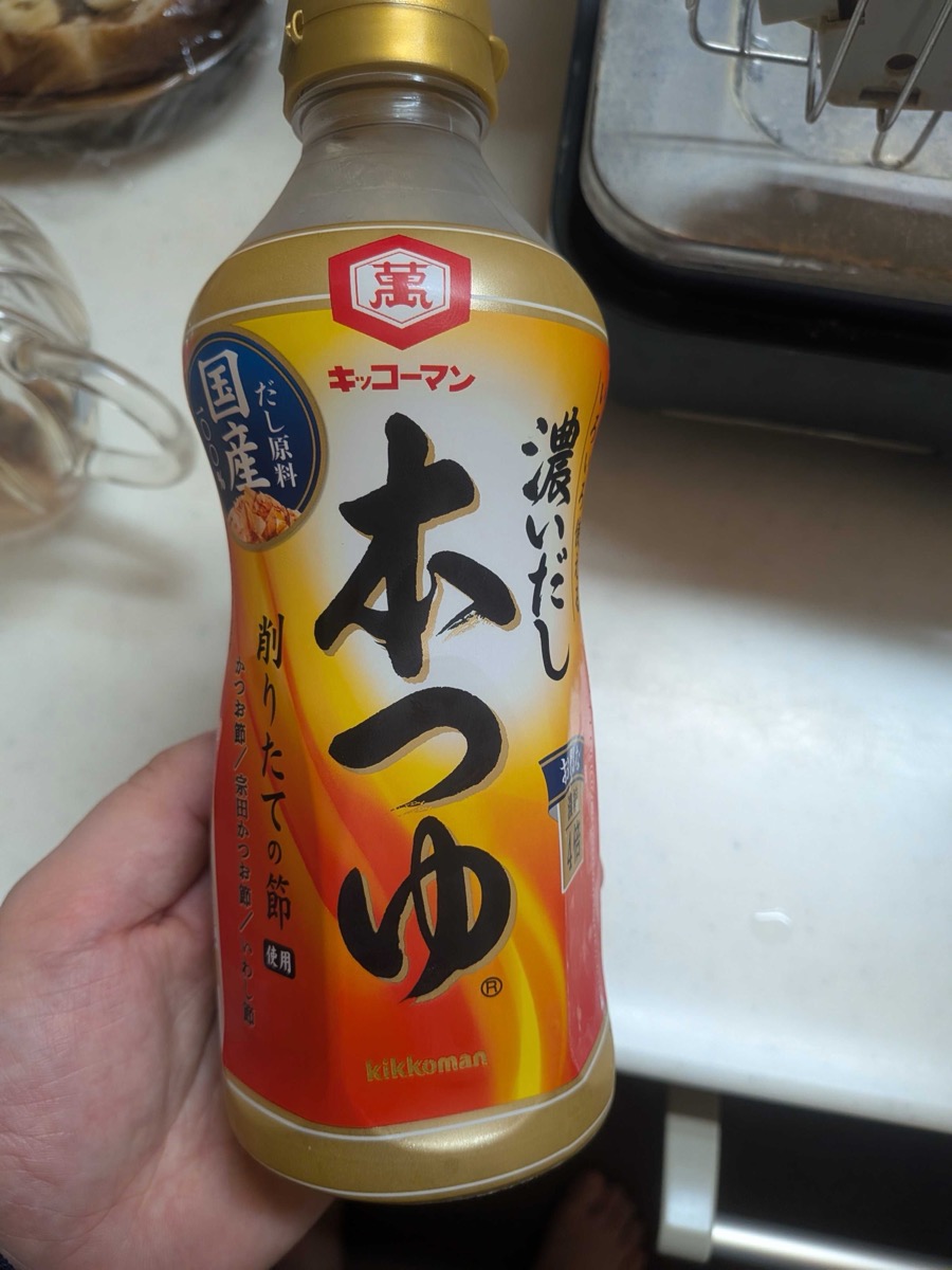 A bottle of Kikkoman Koidashi Hontsuyu — a 4× concentrate Japanese mentsuyu noodle soup base — held in hand on a kitchen counter