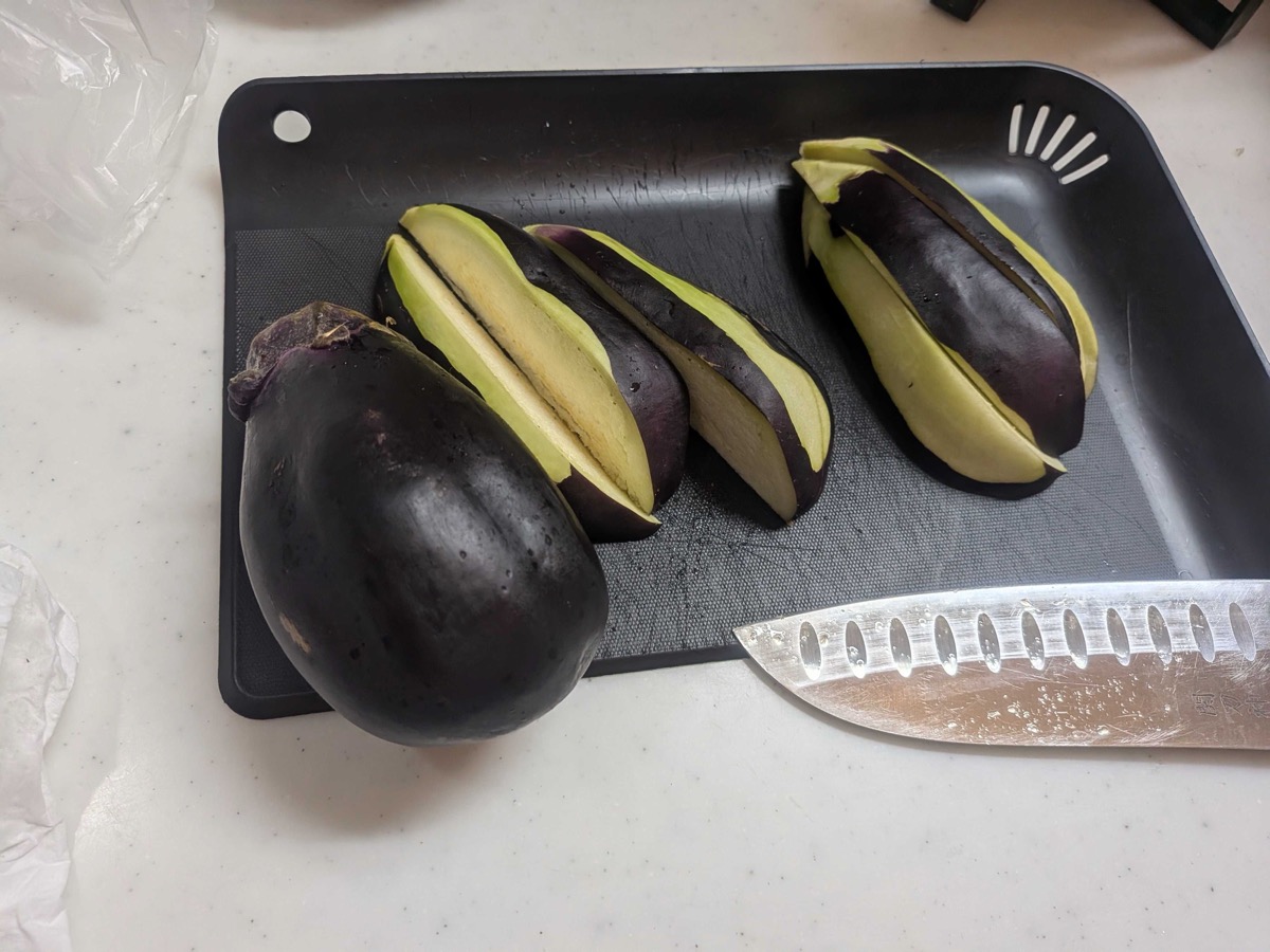 A whole mizu-nasu (water eggplant) on a black cutting board alongside another eggplant cut into wedges with shallow diagonal scoring on the flesh side