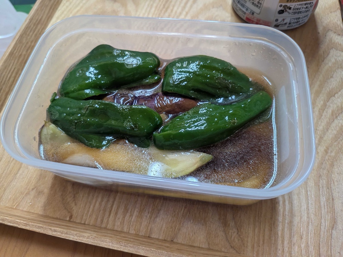 Pan-fried eggplant and green pepper soaking in a clear plastic storage container of dashi-soy tsuyu, ready to chill in the fridge
