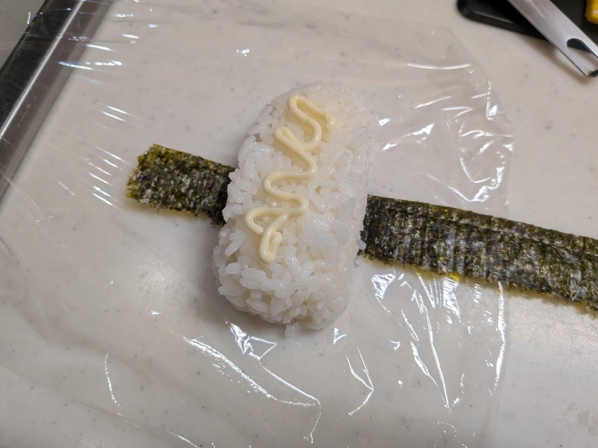 Assembling Spam onigiri — a rice block on a strip of nori on plastic wrap, with a squiggle of Kewpie mayo piped across the top of the rice