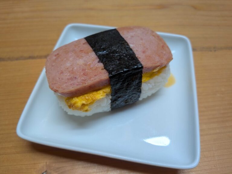 A finished Japanese-style Spam onigiri on a small white square plate, showing layers of rice, sweet tamagoyaki, and a thick slice of pan-fried Spam wrapped with a strip of nori