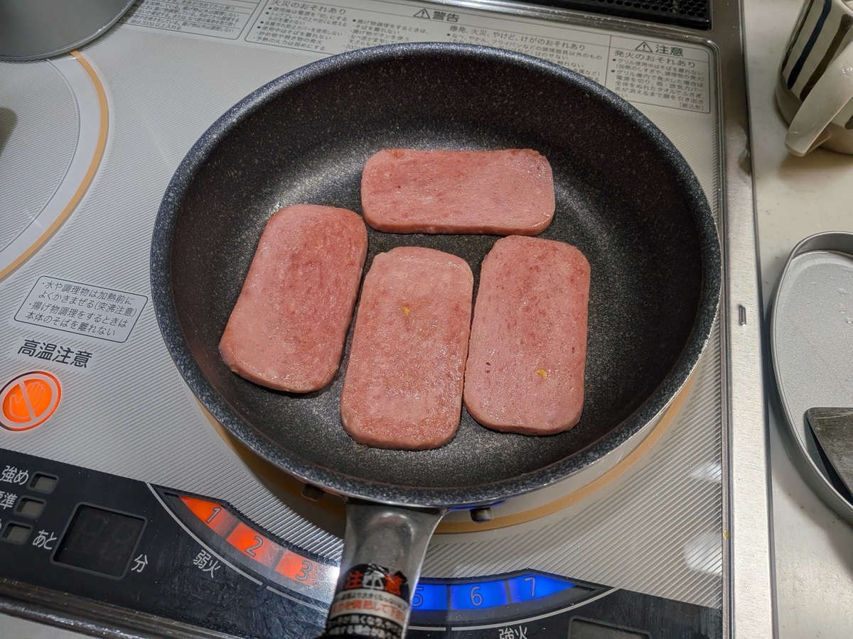 Four slices of pan-fried Spam-style luncheon meat in a black frying pan on an IH cooktop, golden brown on both sides