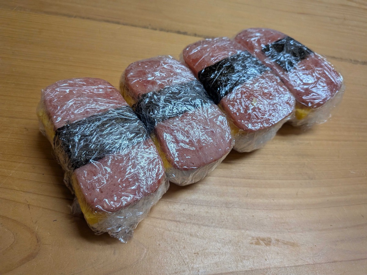 Four assembled Spam onigiri wrapped in plastic wrap, lined up on a wooden cutting board ready to eat or pack into a bento