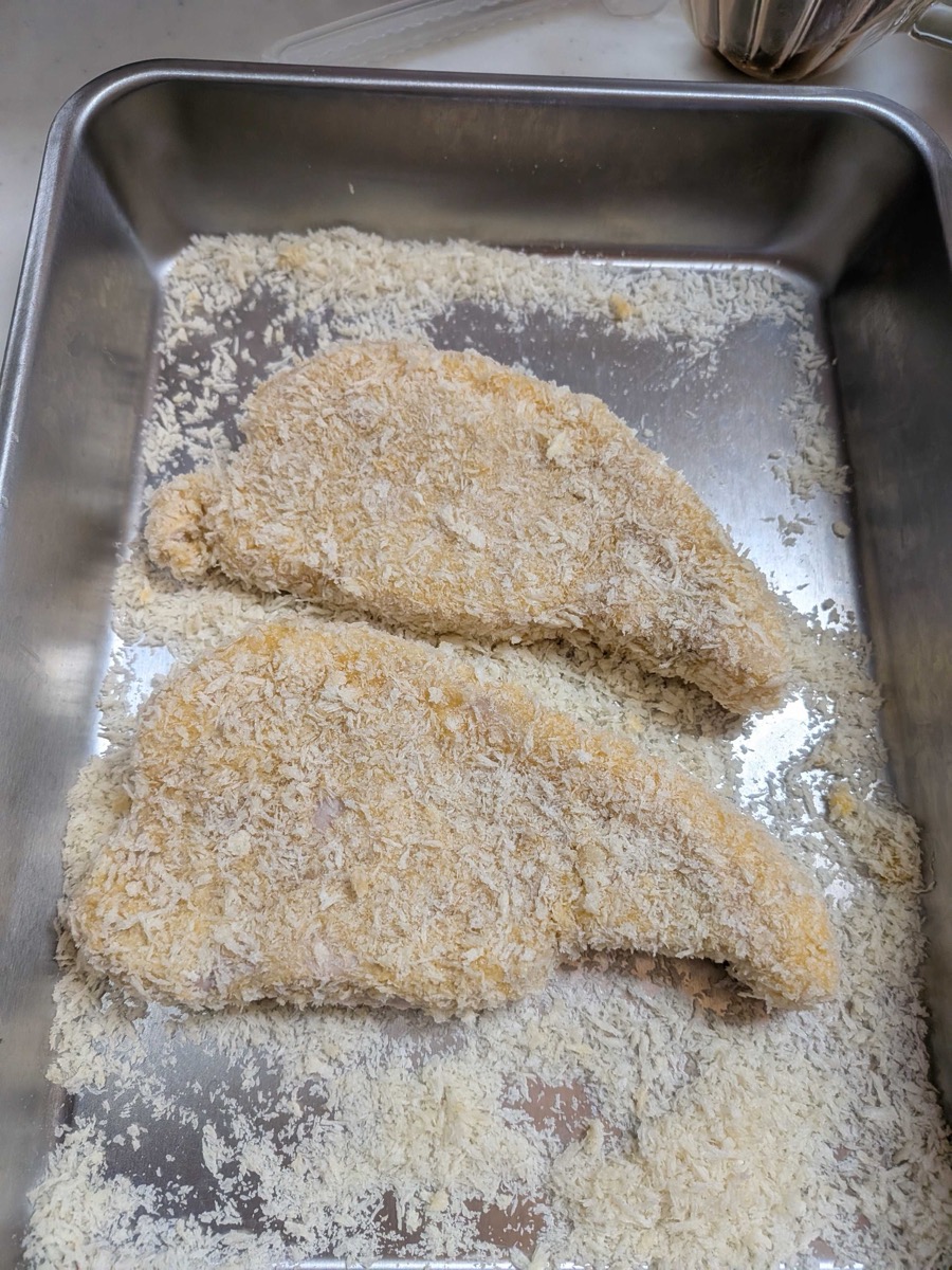 Two tonkatsu pork cutlets fully coated in panko breadcrumbs, resting in a metal tray and ready to deep fry