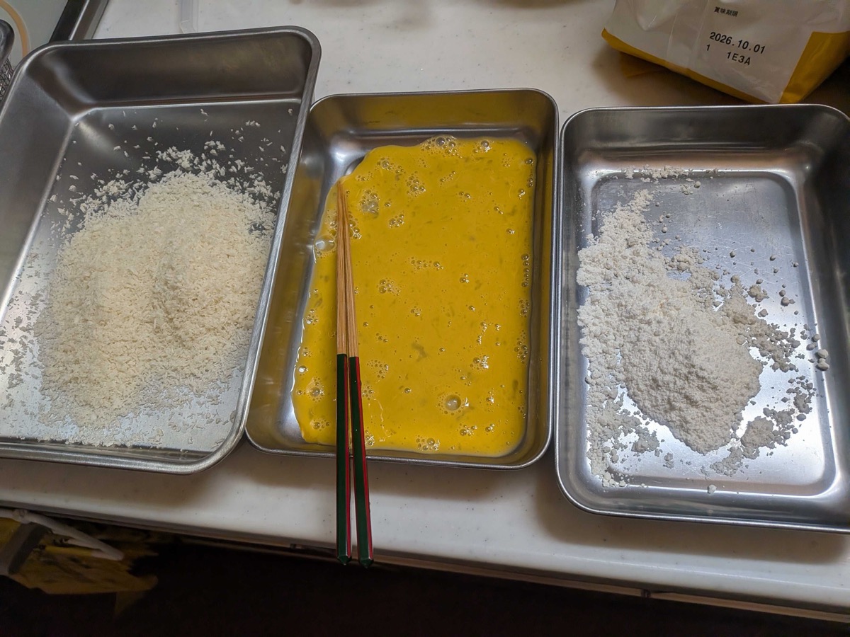 Three metal trays in a row set up as a breading station: panko breadcrumbs on the left, beaten egg in the middle, and all-purpose flour on the right