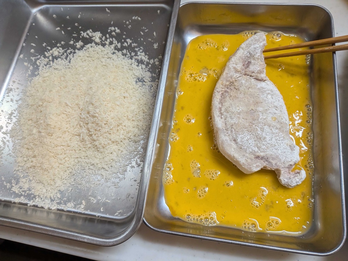 Flour-coated pork cutlet being dipped into beaten egg wash using chopsticks, with panko breadcrumbs tray alongside