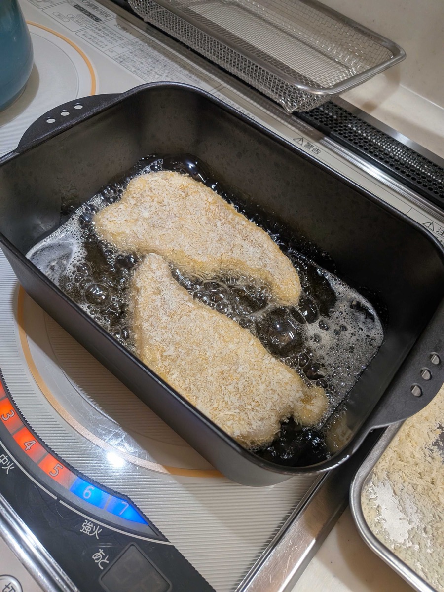 Two breaded tonkatsu cutlets deep frying in hot oil in a black rectangular pan on an IH cooktop, bubbling vigorously