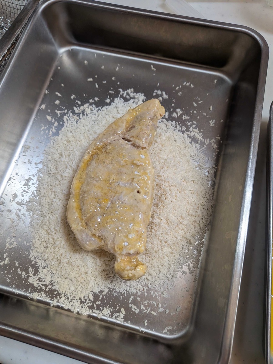 Egg-coated pork cutlet pressed into panko breadcrumbs in a metal tray, being coated on all sides
