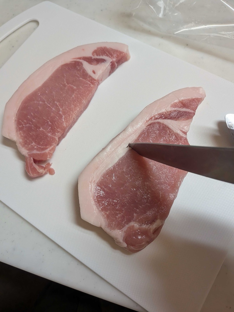 Scoring the fat and connective tissue of pork loin cutlets on a white cutting board with a knife to prevent curling during cooking