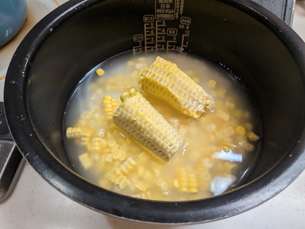Corn kernels scattered over rice with two stripped cobs laid on top in the rice cooker pot, ready to cook