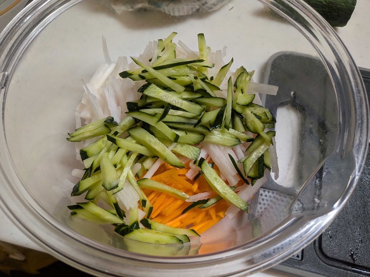Shredded daikon, carrot, and cucumber matchsticks in a glass bowl with a slicer
