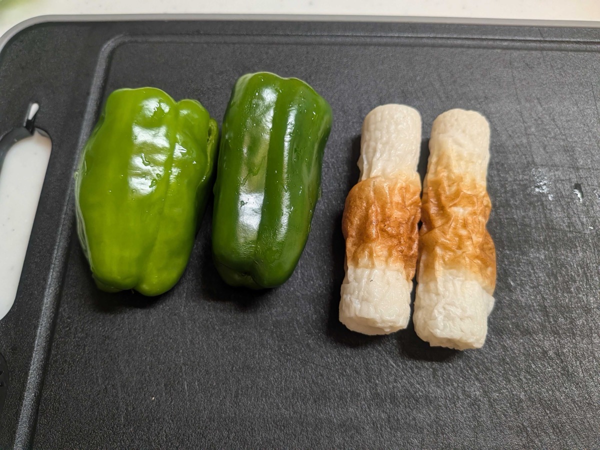 Two whole green peppers and two chikuwa fish cakes on a cutting board