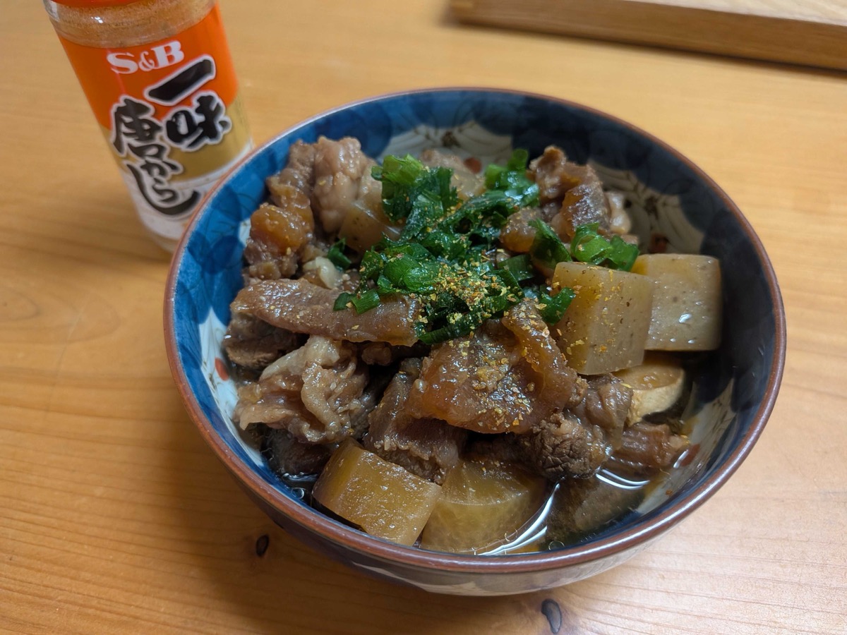 A bowl of finished gyusuji nikomi — Japanese braised beef sinew with cubed daikon and konnyaku in a dark miso broth — garnished with chopped scallion and a dusting of red ichimi togarashi, with an S&B ichimi bottle in the background