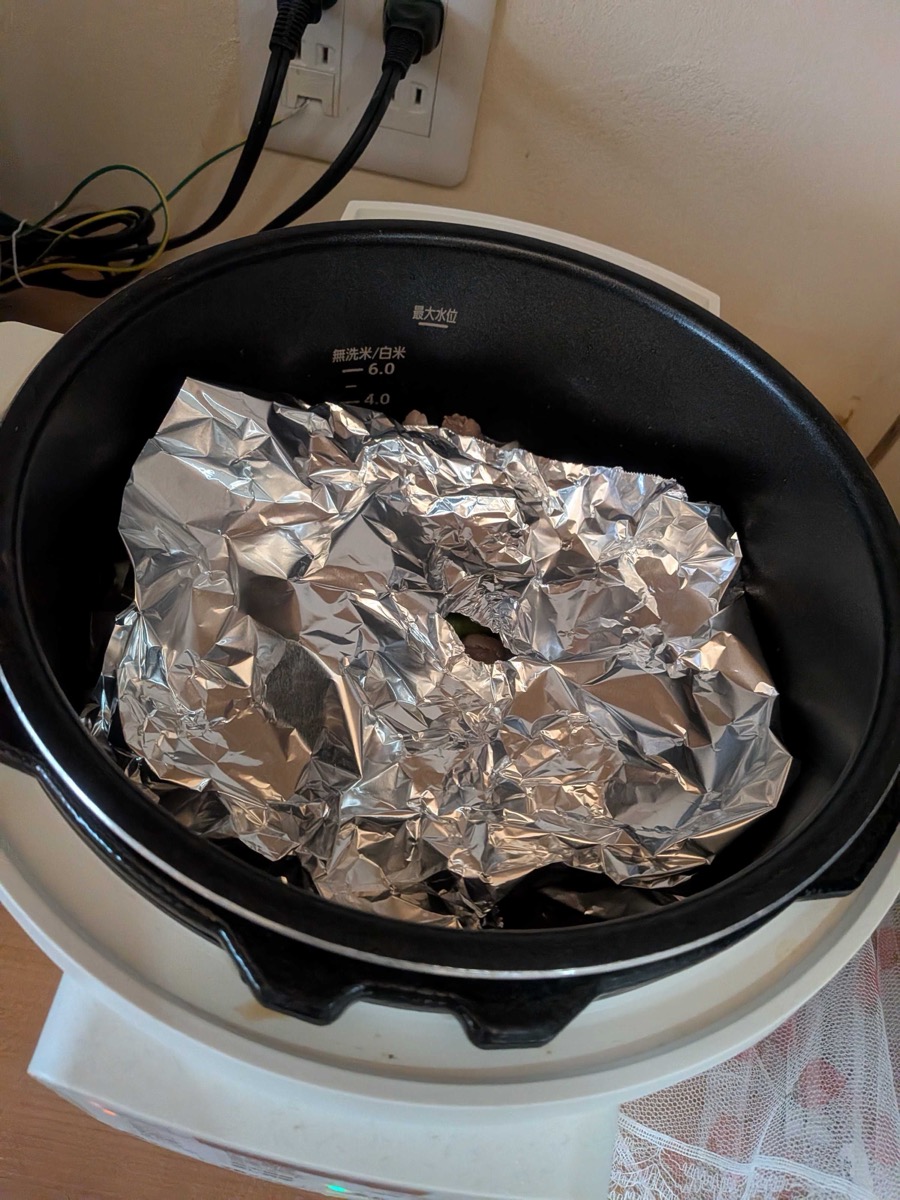 A crumpled sheet of aluminum foil laid directly on the surface of the ingredients in the electric pressure cooker pot — a makeshift otoshibuta drop lid for Japanese gyusuji nikomi
